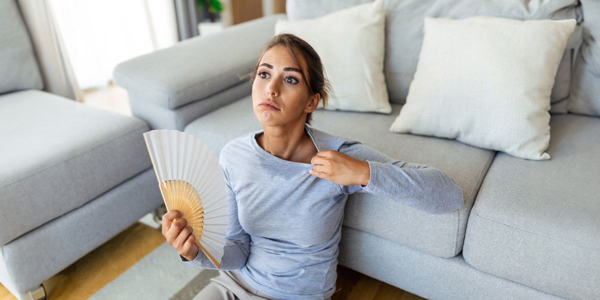 Stressed annoyed woman using waving fan, suffering from overheating. Summer heat health hormone problem. No air conditioner at home. Sat on sofa, feeling exhaustion and dehydration. Heatstroke concept.