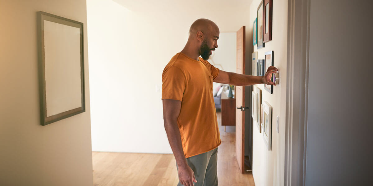 A man in an orange shirt stands in a hallway while adjusting the thermostat.