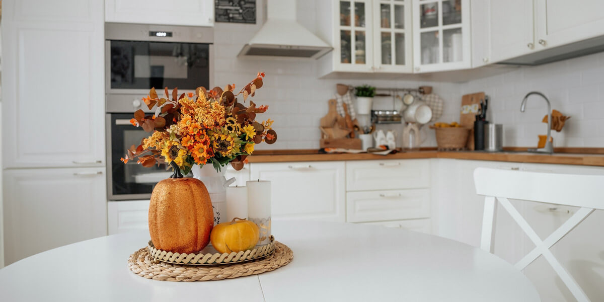 A bright kitchen with white cabinetry and appliances. A round table features a fall-themed centerpiece with flowers, a pumpkin, and a small gourd.