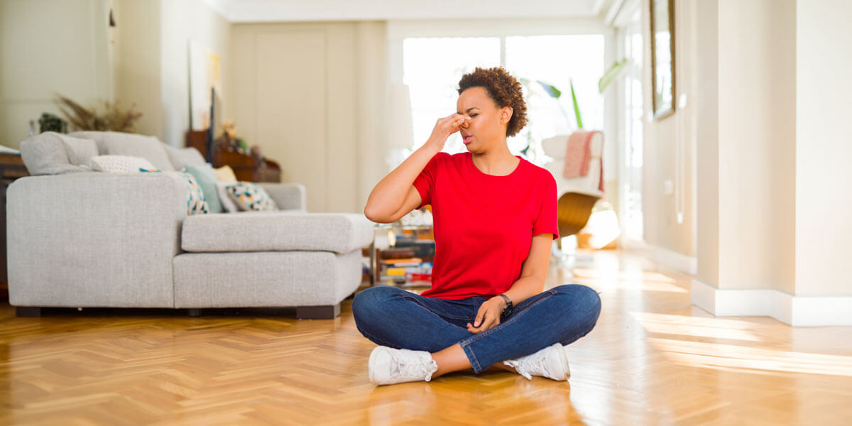 Person sitting cross-legged on the floor in a well-lit living room, practicing a breathing exercise with one hand pinching their nose.