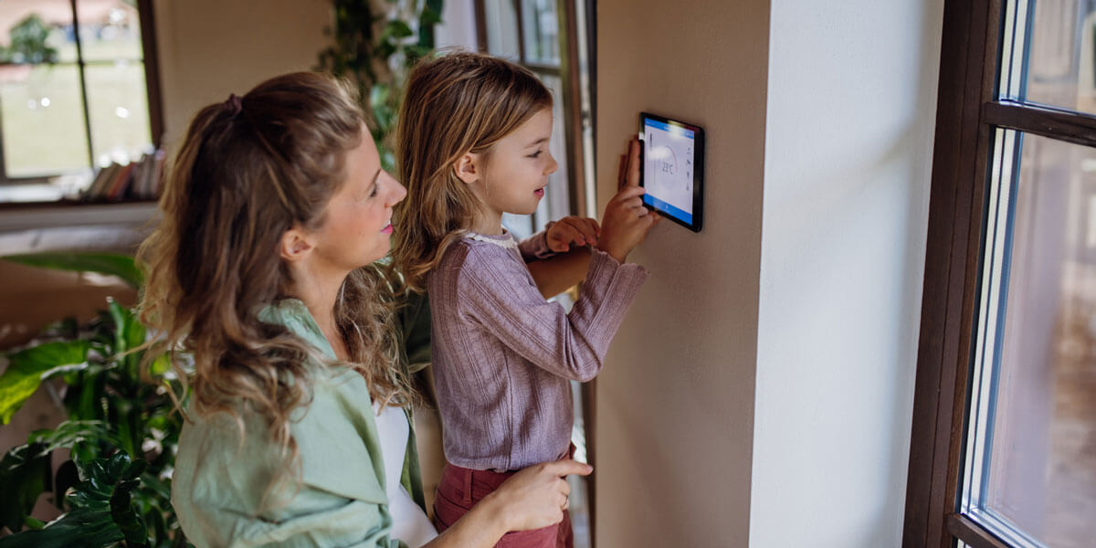 A woman and a young girl interact with a wall-mounted smart home control panel in a bright, cozy home setting.